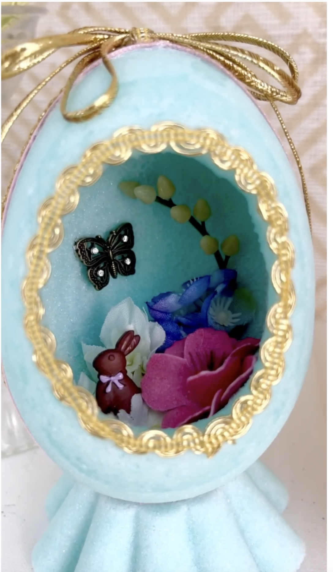Panoramic Sugar Easter Eggs. A Tiffany Blue sugar egg shell decorated with diorama scene inside with a tiney butterfly and chollate Easter bunny nested in a bed for pink, blue and white flowers, the outside is trimmed in gold ribbon with adorning the outside of the egg.